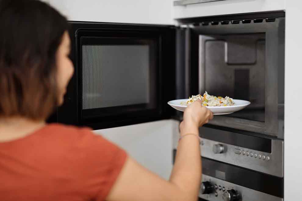 requentar comida  mulher esquentando um prato de comida em um microondas preto em uma cozinha clara
